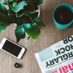 Flat lay arrangement of a coffee mug, newspaper, smartphone, and plant on a wooden table.